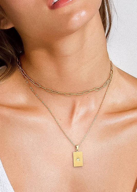 Close-up of a person wearing a gold necklace with a rectangular pendant on a plain background