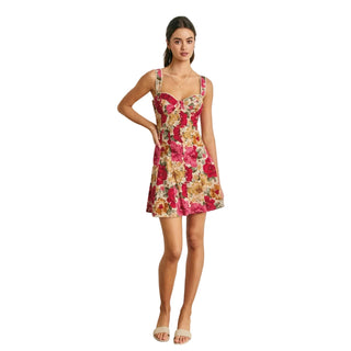 A woman modeling a sleeveless floral mini dress with a sweetheart neckline and wide shoulder straps, standing against a white paneled wall.