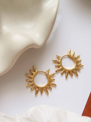 A pair of gold-toned sunburst hoop earrings displayed on a white surface next to a beige ceramic dish.