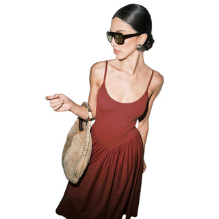 A woman wearing a rust-red spaghetti-strap midi dress, black sunglasses, and a beige suede shoulder bag, posing against a plain gray wall.