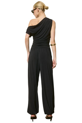A woman modeling a black one-shoulder draped jumpsuit from the back, standing against a plain light gray wall.