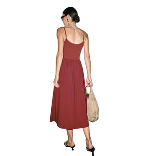 A woman wearing a rust-red spaghetti-strap midi dress, viewed from behind, holding a beige tote bag against a plain light gray wall.