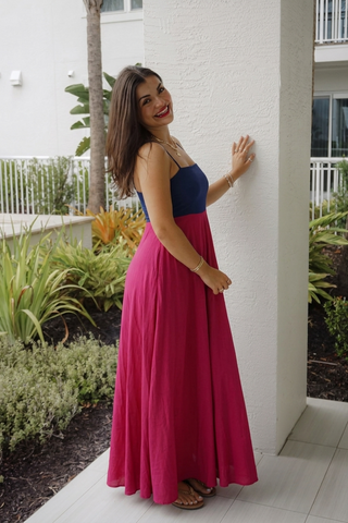 A woman wearing a two-tone maxi dress with a navy blue bodice and a vibrant magenta pink skirt, posing outdoors against a white stucco wall.