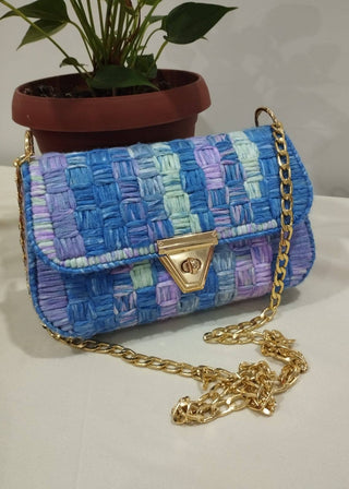 Blue woven handbag with gold chain strap on a light surface with a plant in the background