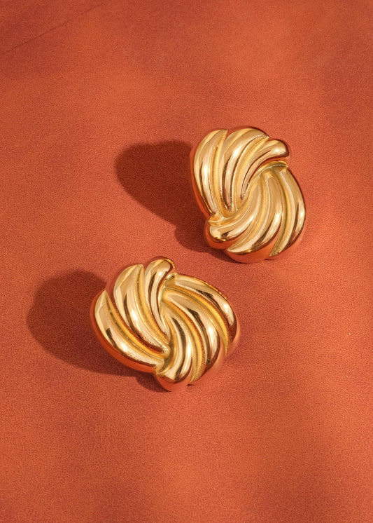 A pair of gold-colored wavy rectangular stud earrings displayed on a brown surface.