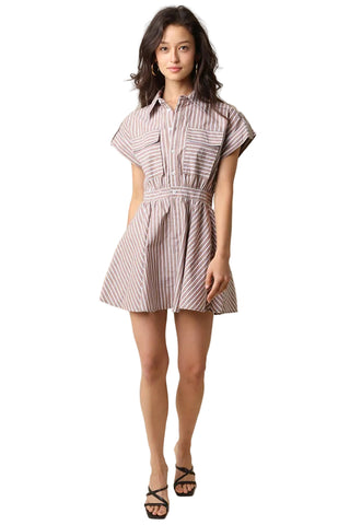 A woman modeling a short-sleeve collared shirt dress with vertical pink and white stripes, featuring a button-up front, chest pockets, and a gathered waist, paired with black strappy sandals against a solid beige background.