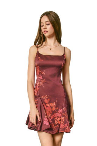 A woman modeling a satin slip dress with a floral print in deep burgundy and pink, set against a plain light gray studio background.