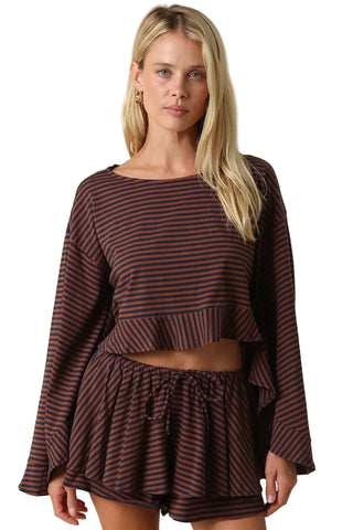 A woman modeling a matching two-piece lounge set featuring a dark brown and black striped long-sleeve crop top with a ruffle hem and matching drawstring shorts, set against a solid beige background.
