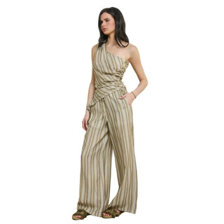 A woman models a one-shoulder striped jumpsuit with wide legs and a gathered waist, paired with brown sandals, standing against a textured beige wall.