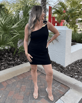 Woman in a black one-shoulder dress standing outdoors with plants and a building in the background.