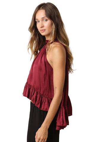 A woman wearing a sleeveless halter-neck top in deep burgundy with a ruffled hem, paired with black pants, photographed against a solid light beige background.
