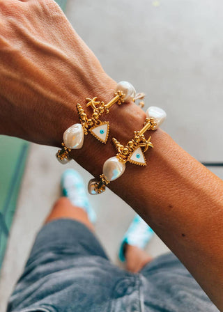 A person's wrist wearing a gold-plated bracelet adorned with white pearl-like beads and small gold animal eye-shaped charms.