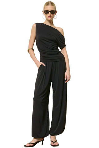 A woman modeling a black one-shoulder draped jumpsuit with wide-leg pants, styled with black sunglasses, hoop earrings, a gold bracelet, and black heeled sandals against a plain light gray wall.