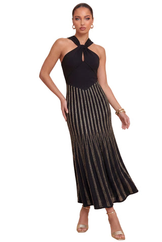 A woman modeling a sleeveless halter-neck maxi dress with a black bodice and a pleated, metallic gold-and-black striped skirt, set against a solid beige studio background.