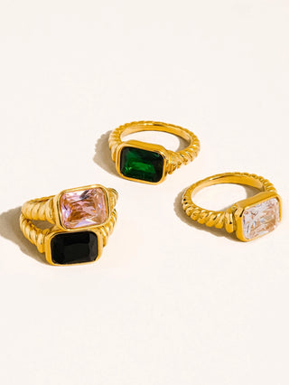 Three gold rings with rectangular gemstone settings arranged on a light cream background. One ring has a green stone, one has a black stone, and one has a pale pink stone.